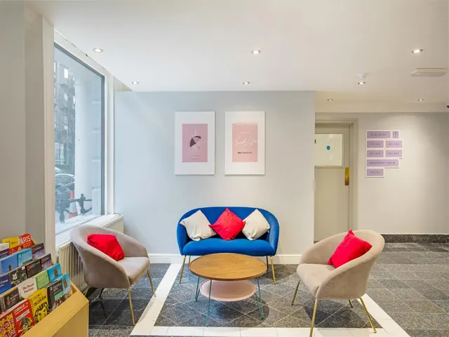 A lobby seating area with a blue two-seat sofa, two beige armchairs with red cushions, a round wooden coffee table, and a book rack beside a window onto the street.
