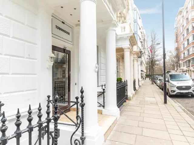 The white stucco facade of a townhouse hotel with a Townhouse sign, columns and black iron railings, beside a street with parked cars and a white van.