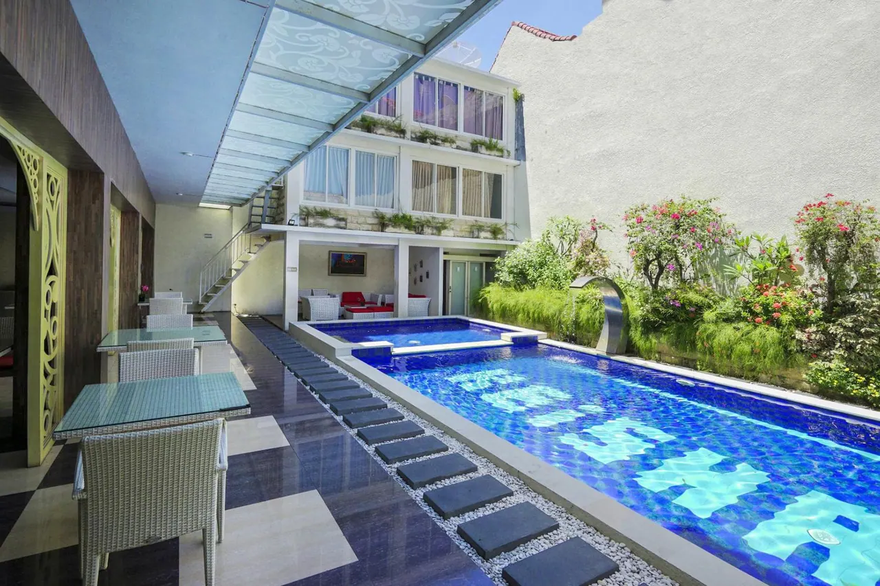 A long blue-tiled swimming pool with stepping stones over white gravel runs beside a covered dining area, with a smaller pool and planted garden beyond.