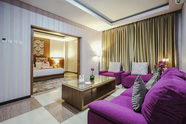 A living area with purple sofa and armchairs, a glass coffee table and gold curtains, with a bedroom visible through a sliding door.