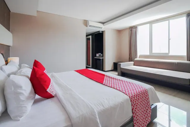A bedroom with a made double bed with white linens, red and white pillows and a red patterned runner, a grey sofa beneath a large window, and a wall-mounted air conditioner.