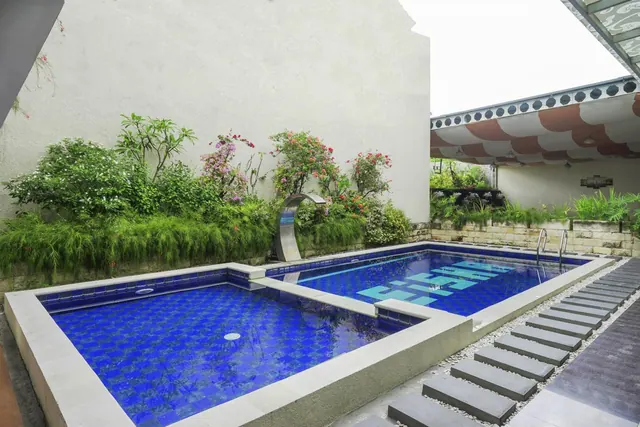 A blue-tiled pool with a stainless steel water feature, flowering shrubs and a striped covered pavilion beside it.