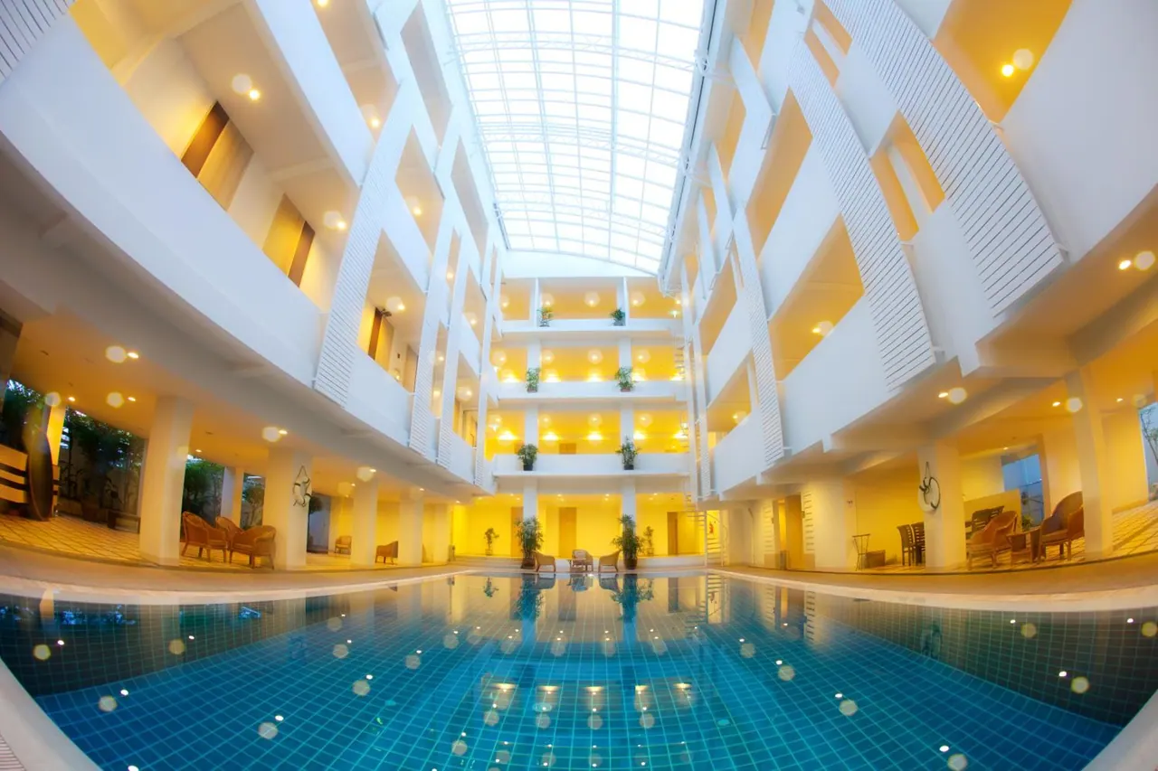 An indoor swimming pool sits beneath a glass skylight, surrounded by multi-level white balconies and warmly lit atrium walkways.