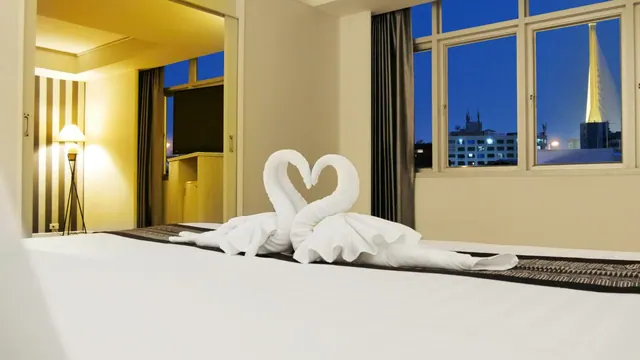 A made bed with two white towel swans arranged head to head, beside a window showing city buildings at dusk.