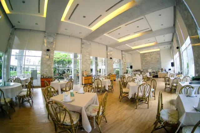 A wide-angle view of a dining room with white-clothed tables, rattan chairs with floral cushions, and tall windows showing trees outside.