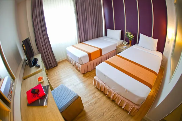 A bedroom with two single beds with orange and white bedding, a purple feature wall, a wall-mounted television on a wooden desk, and a window with sheer and purple curtains.