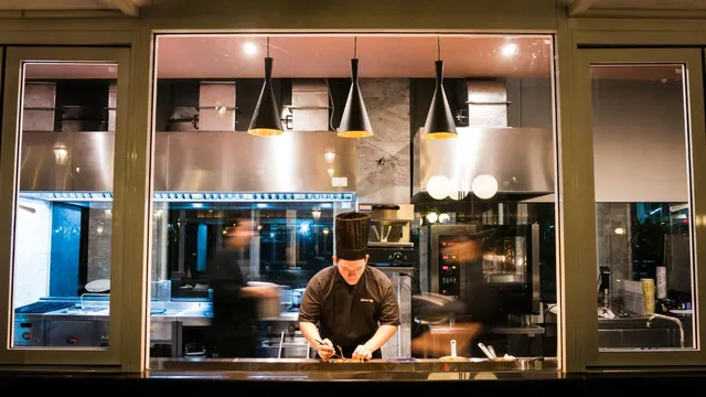 A chef in a tall toque prepares food at an open kitchen counter seen through a large glass window, lit by black pendant lamps.