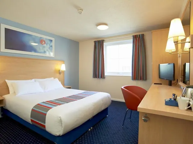 A double bed with a striped runner stands beside a window, with a desk, television and kettle on the right.
