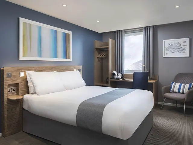 A double bed with white bedding and a grey runner sits beside a desk, an armchair and a wardrobe, with a window looking onto a building facade.