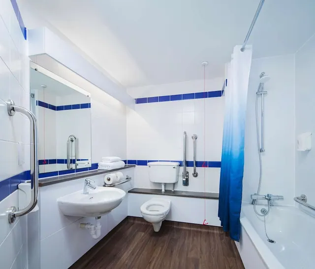 A bathroom with a wall-mounted basin, a toilet with grab rails, and a bath with an over-bath shower and blue shower curtain.