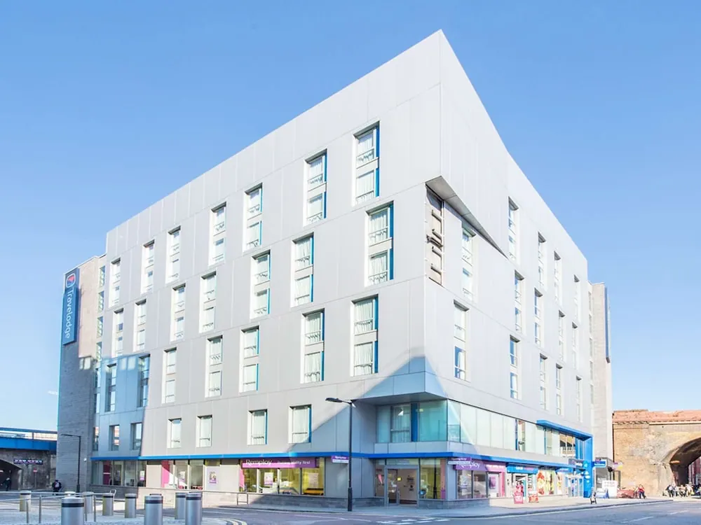The exterior of a white modern Travelodge hotel building with blue accents under a clear blue sky, with bollards and a street in the foreground.