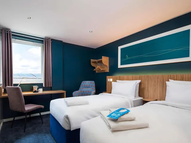 A twin hotel bedroom with two made beds, a desk and upholstered chair by the window, and a kettle and mugs on the desk.