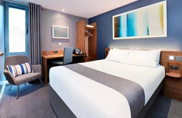 A hotel bedroom with a double bed, a wooden desk and chair, an armchair by the window, and an open wardrobe, with a large abstract painting above the bed.