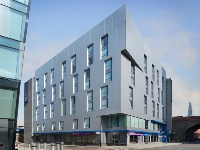 The exterior of a grey-clad multi-storey Travelodge hotel on a street corner, with The Shard visible in the background on the right.