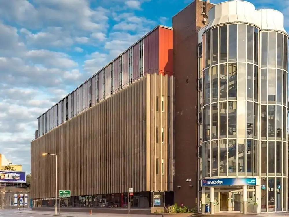 Exterior of a Travelodge hotel building with curved glass towers and a slatted facade under a partly cloudy sky, with the hotel entrance sign visible at street level.