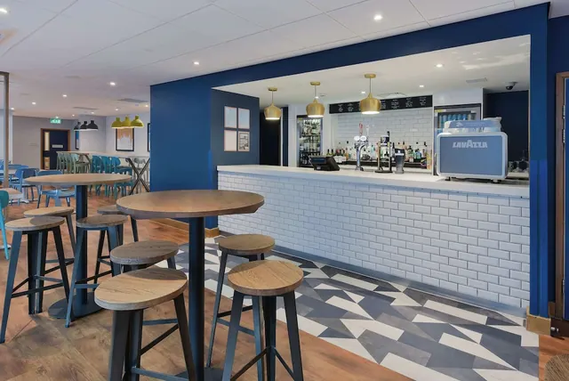 A bar area with high wooden tables and stools, a white tiled bar counter with taps, and a Lavazza coffee machine on the right.