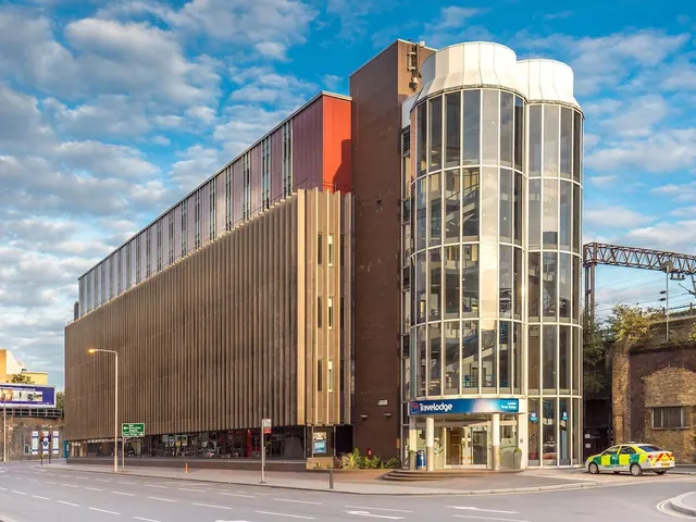 The exterior of a multi-storey Travelodge hotel with a glass cylindrical tower, a blue entrance canopy and a green and yellow car parked on the street beside it.