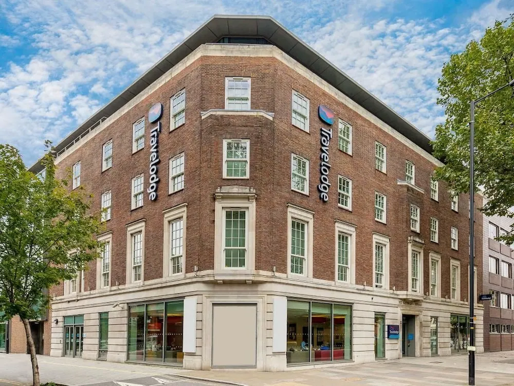 A brick corner building with Travelodge signage and ground-floor shopfronts stands on a street under a partly cloudy sky.