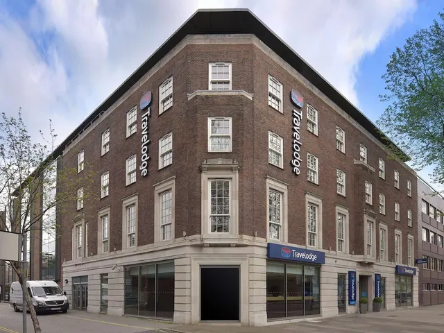 Exterior of a brick corner building with Travelodge signage, seen from the street, with a white van parked at the left edge.