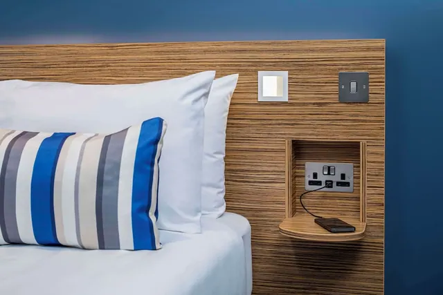 A made bed with white pillows and a striped cushion sits beside a wood-panelled headboard with wall-mounted sockets and a shelf where a phone is charging.