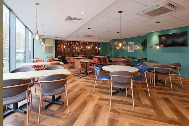 A bright dining room with herringbone wood flooring, grey and blue chairs at round and rectangular tables, teal walls, a wall-mounted television and large windows on the left.
