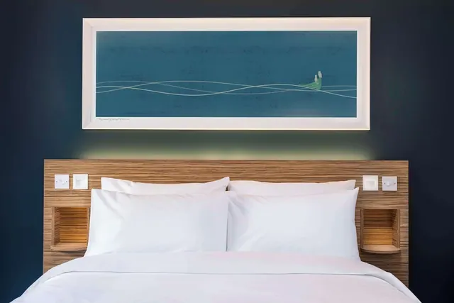A made double bed with white pillows sits against a wooden headboard below a framed blue abstract artwork on a dark navy wall.