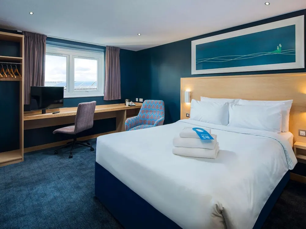 A made double bed with folded towels and a blue door hanger sits beside a desk, armchair and wardrobe in a room with dark blue walls.