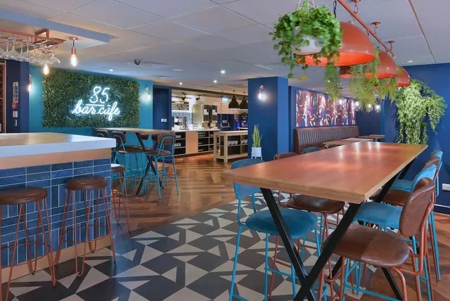 A bar cafe area with a lit neon sign reading 85 bar cafe, a green plant wall, a long wooden table with teal chairs, high stools at a counter, and hanging plants under orange pendant lamps.