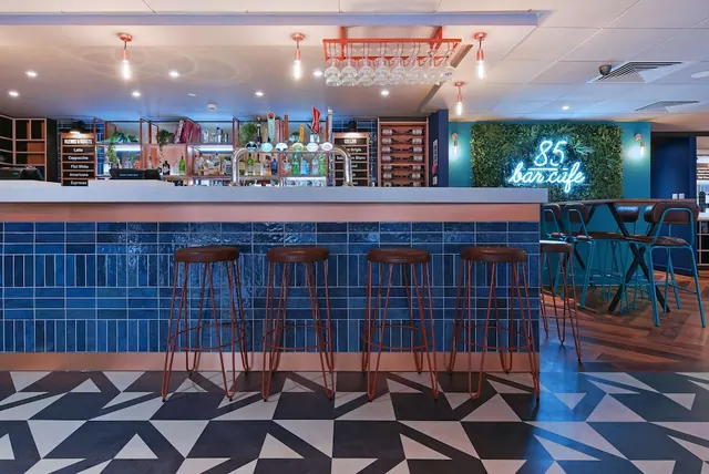 A bar counter with a blue tiled front, four brown bar stools, and a neon sign reading 85 bar cafe in front of a green living wall.