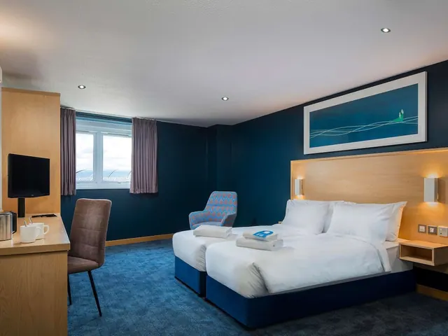 A hotel bedroom with two beds, a blue patterned armchair, a desk with a television and kettle, and dark blue walls.