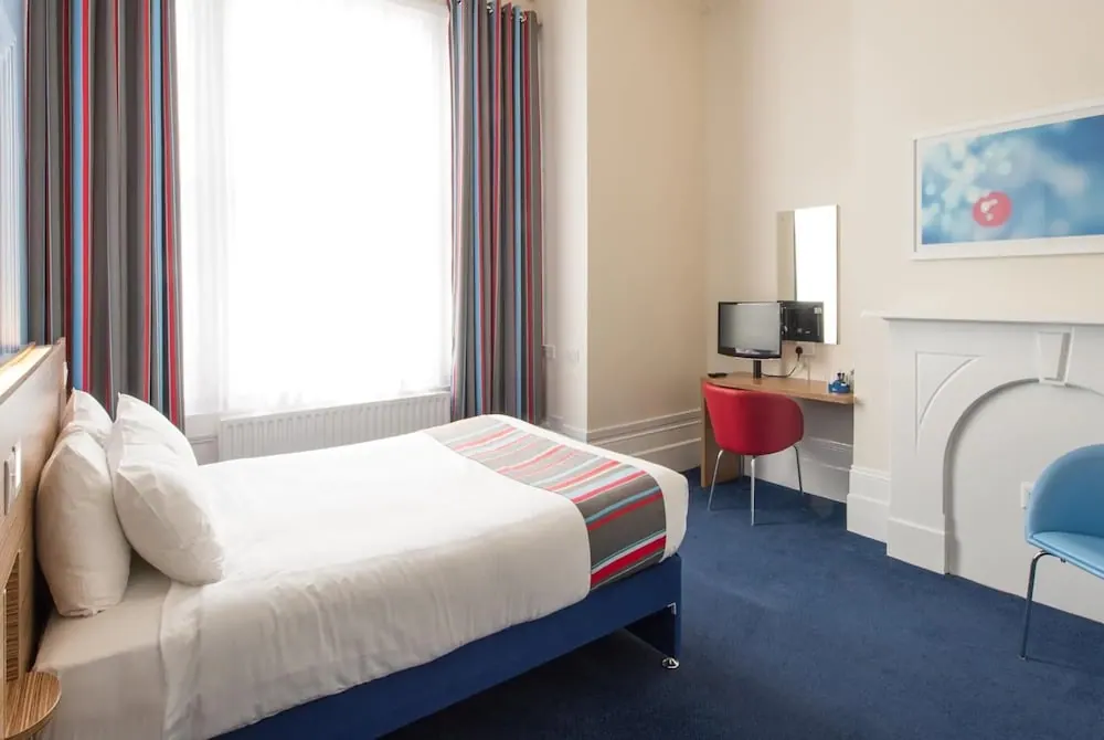 A double bed with a striped throw sits beside a bright window, with a desk, red chair and monitor in the corner.