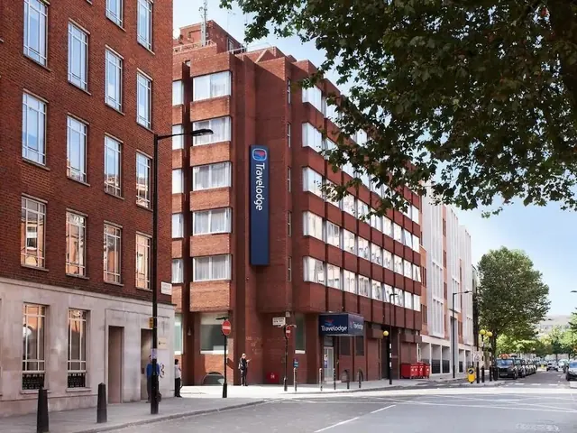 Exterior of a red brick Travelodge hotel building on a city street, with a tall blue sign on its facade and a tree in the foreground.