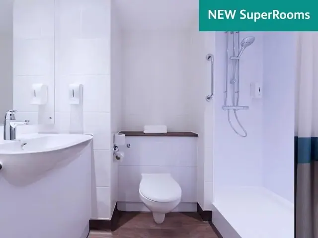 A white bathroom with a wall-mounted toilet, a single wash basin with mirror, a walk-in shower with a grab rail, and a teal banner reading NEW SuperRooms in the corner.