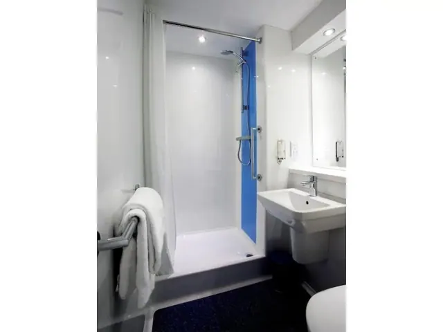 A white bathroom with a walk-in shower behind a curtain, a blue wall panel, a grab rail holding towels, and a pedestal sink beneath a mirror.