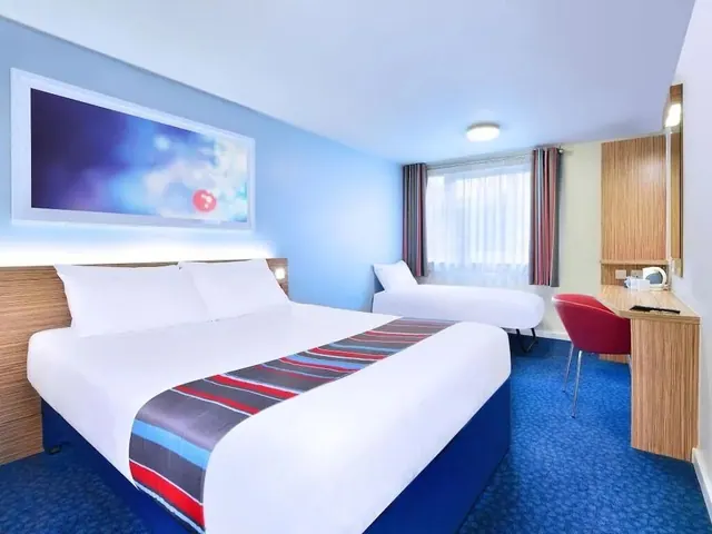 A hotel bedroom with a double bed, a single bed by the window, and a wooden desk with a red chair.