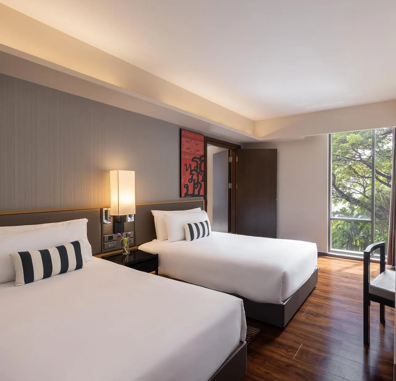 A bedroom with two made-up double beds with white bedding, a lit bedside lamp, a red wall artwork, and a tall window looking onto trees.