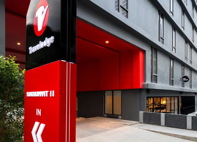 Exterior of a grey multi-storey hotel building with a red entrance canopy and a tall red Travelodge Sukhumvit II sign pillar.