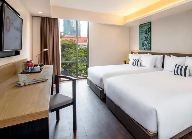 A hotel room with two made-up double beds with white linen, a work desk with a kettle and tray, a wall-mounted television, and a tall window looking out onto trees and a city tower.