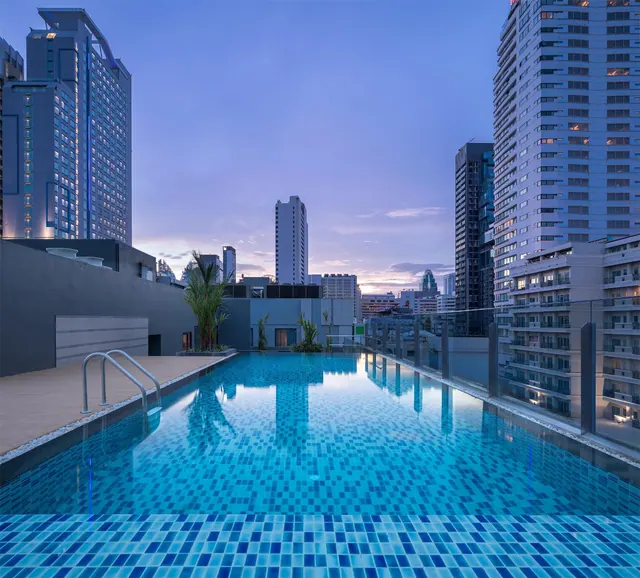 A rooftop swimming pool at dusk with high-rise buildings of the city rising behind it.