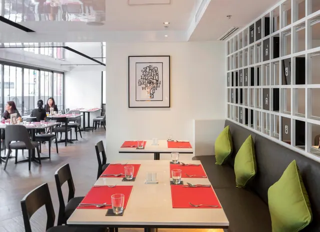 A restaurant dining room with set tables, red placemats, a banquette with green cushions, framed wall art, and a few guests seated near the windows.