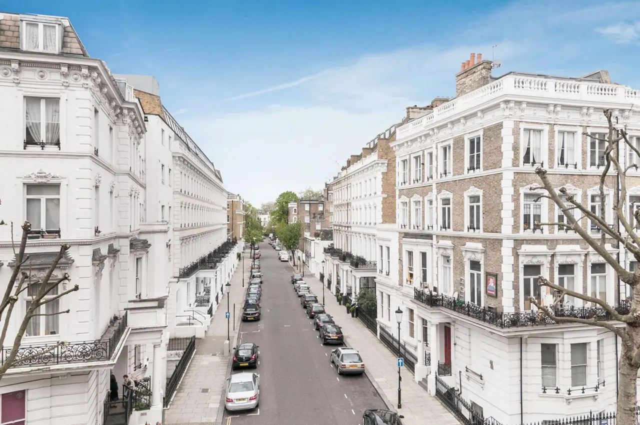 A view down a residential street lined with white and brick Victorian terraced houses and parked cars, taken from an upper floor.