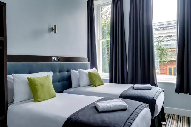 Two single beds with grey throws, folded white towels and green cushions in a bedroom with navy curtains and windows looking onto trees and a brick building.