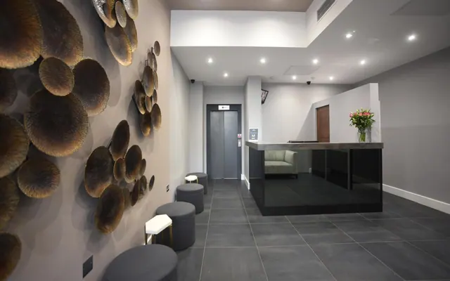 A hotel lobby with bronze disc wall decorations, grey stools, a lift, and a black reception desk holding a vase of flowers.