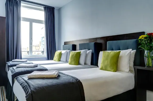 A bedroom with three single beds, green cushions and folded towels, with a window looking out over city buildings.