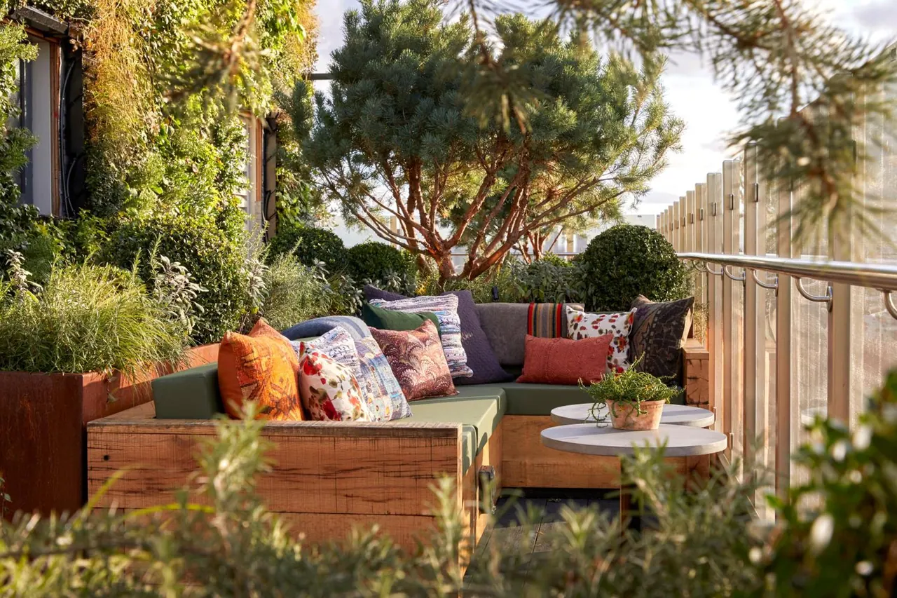 A wooden corner banquette with green cushions and colourful throw pillows sits on a planted rooftop terrace beside round white side tables.