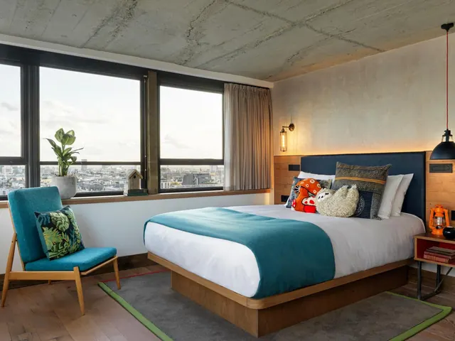A bedroom with a double bed, a teal armchair beside tall windows overlooking city rooftops.