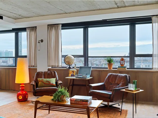 A living area with two leather armchairs, a wooden coffee table, a record player and a large window overlooking the city.