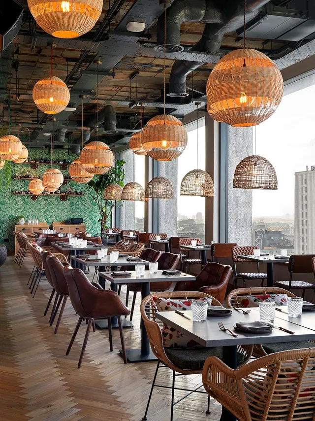 A dining room with wicker pendant lights, leather chairs and set tables, with floor-to-ceiling windows looking out over city buildings.