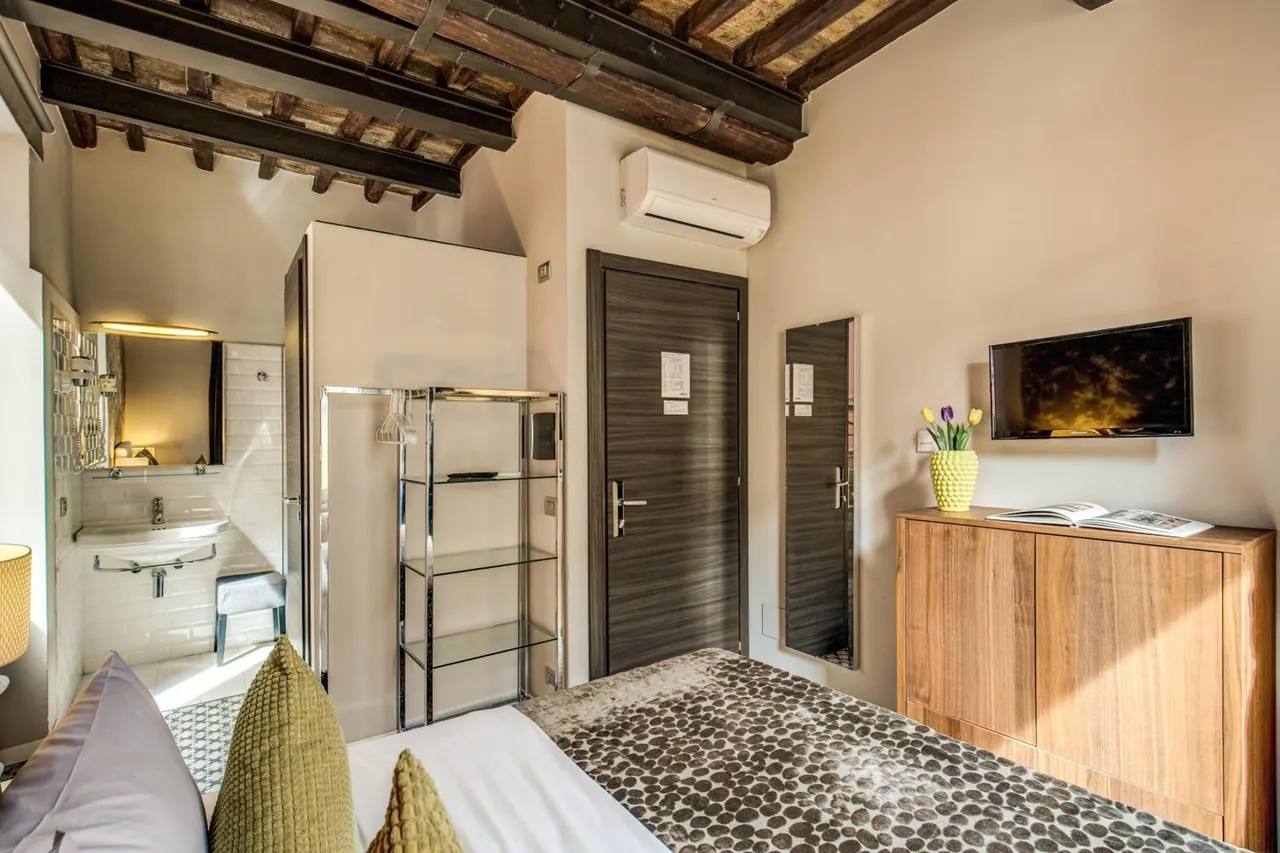 A bedroom with an exposed wooden beamed ceiling, a bed with green cushions, a wall-mounted television, a wooden cabinet, and a bathroom with a sink visible on the left.
