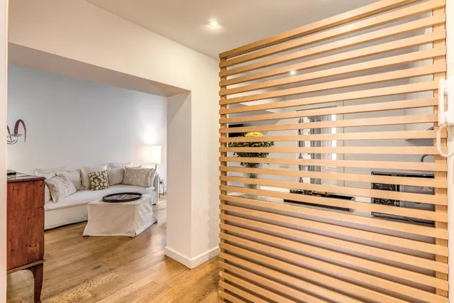 A wooden slatted divider stands in a hallway, with a living room containing a white sofa and ottoman visible beyond.